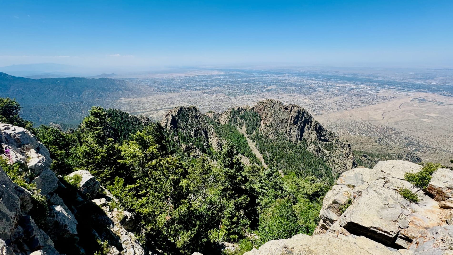 View from a rocky mountain ridge, overlooking a shorter rocky ridge in the midground, and a wide desert grassland valley below. Skies are mostly clear with just a few distant clouds. Haze is visible as a result of wildland fires in the region.