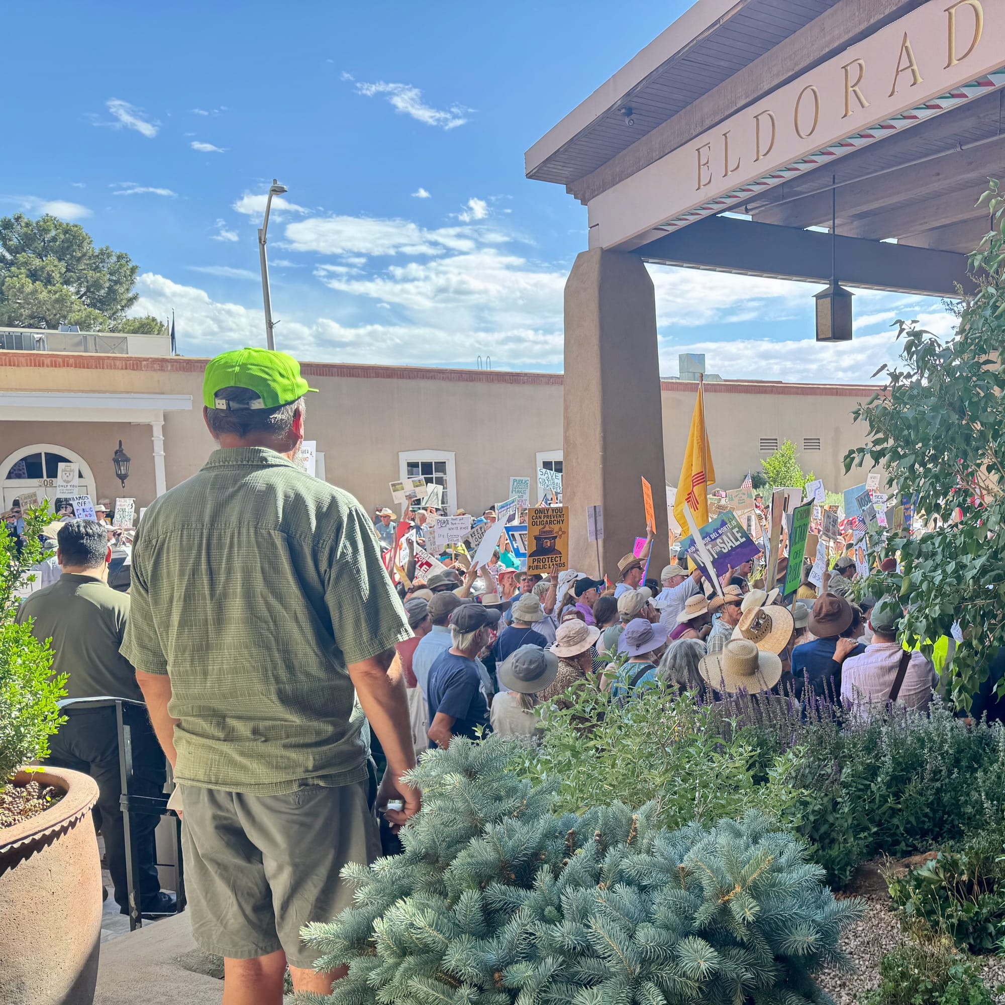 A crowd of people holding protest signs stands in front of a stucco / adobe building and under the roof of a hotel drive-through area. One observer in a bright green hat is present and facing the crowd, which is loud but peaceful. Mostly clear skies are overhead.