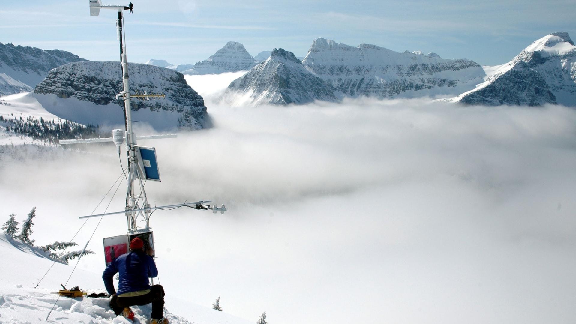A weather station being examined by a person on the side of a snowy mountain with clouds below and mountains peeking out behind.