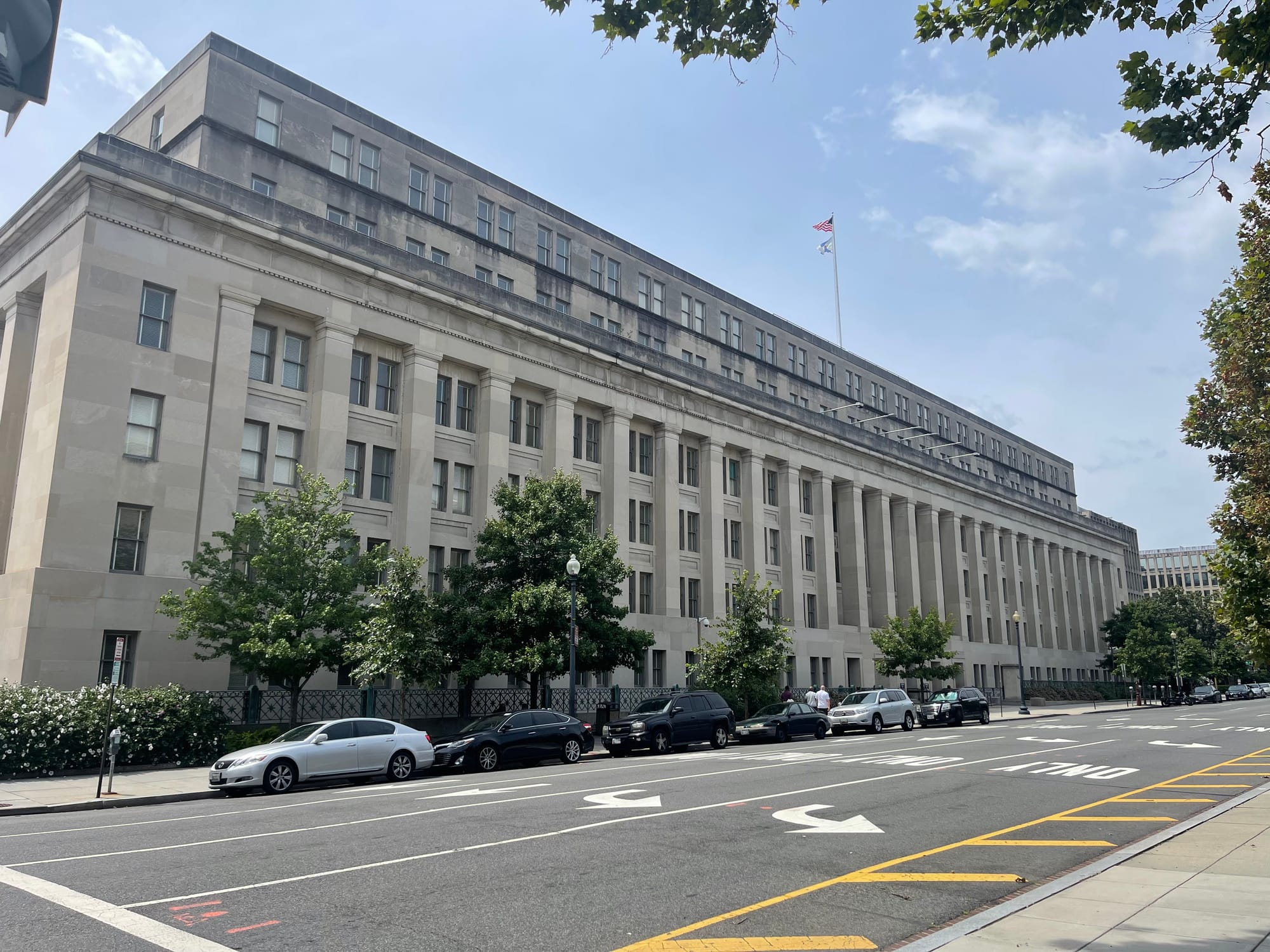 A large stone building showing six stories rises above the street, which has six cars parked along its margin in the foreground. Flags are flying above the building, under mostly clear skies.