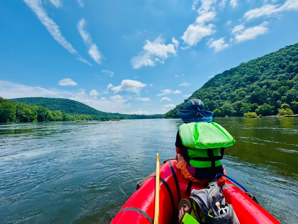 A young person sits on the bow of a red packraft in the middle of a river, with tree-covered hills along the sides of the river and mostly clear skies overhead.