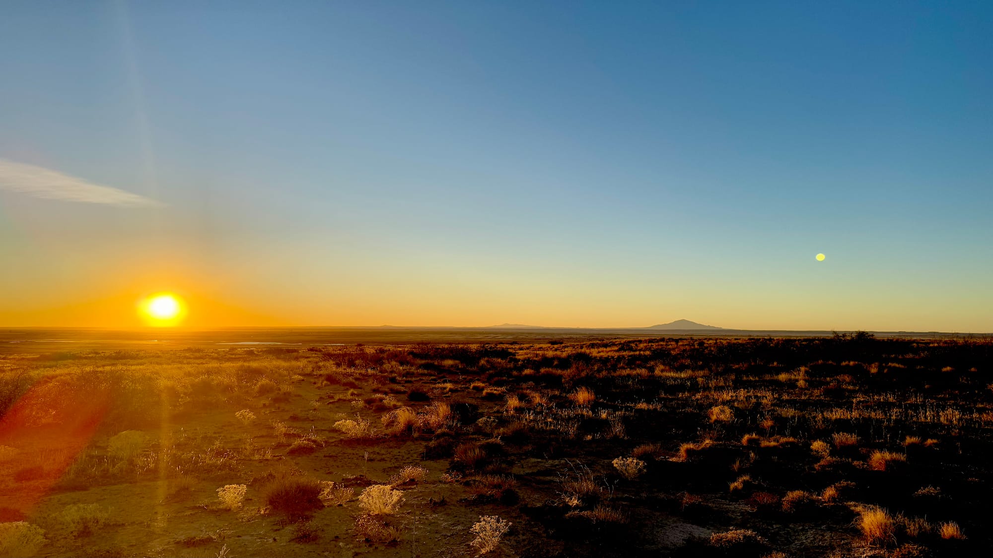 Sunset over desert grassland under clear skies with a few mountains in the distance.