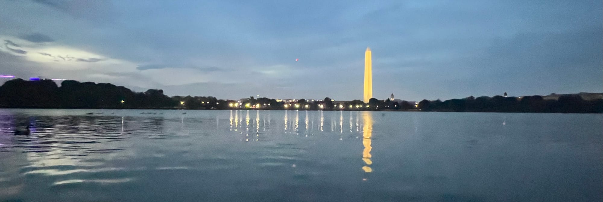 A body of water is in the foreground reflecting lights on the horizon, including a well-lit, off-white obelisk (the Washington Monument) under cloudy skies.