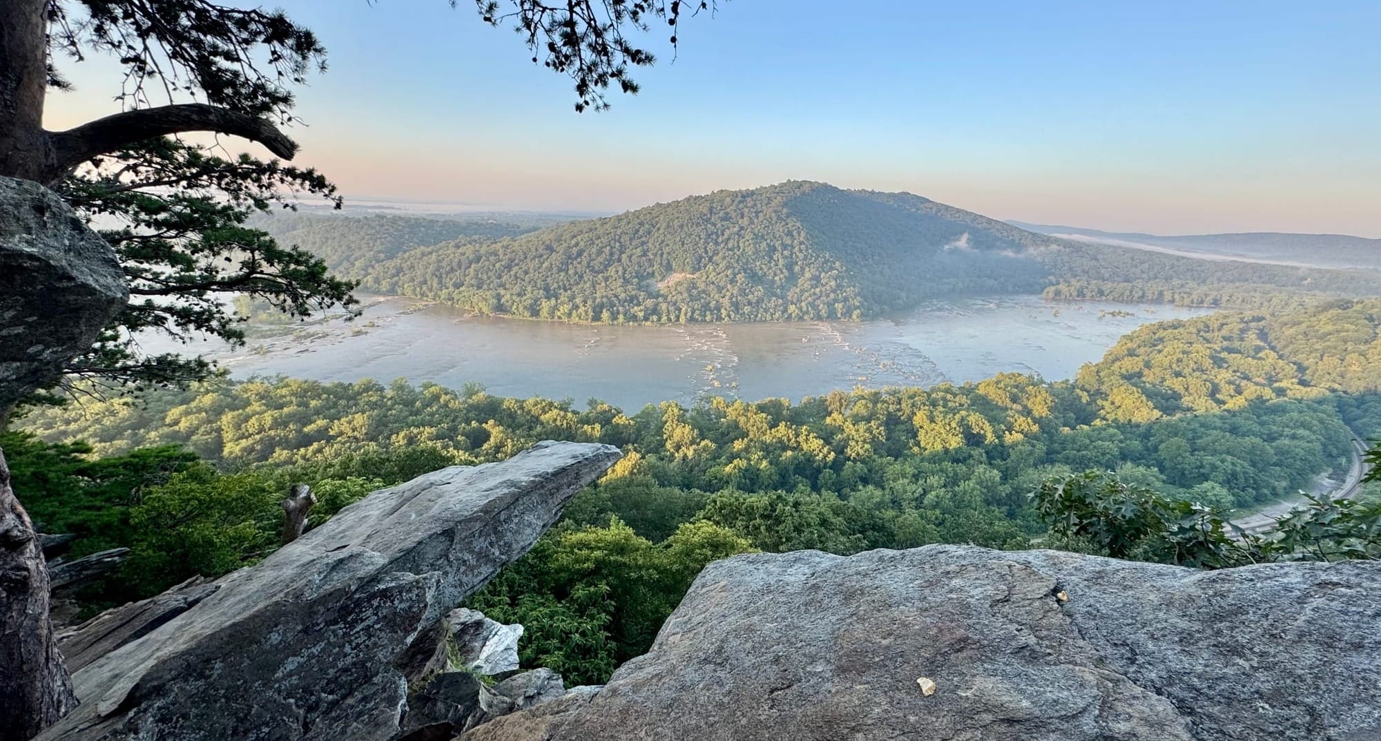 View from a rocky outcrop overlooking a river with wooded hills all around, under clear skies.