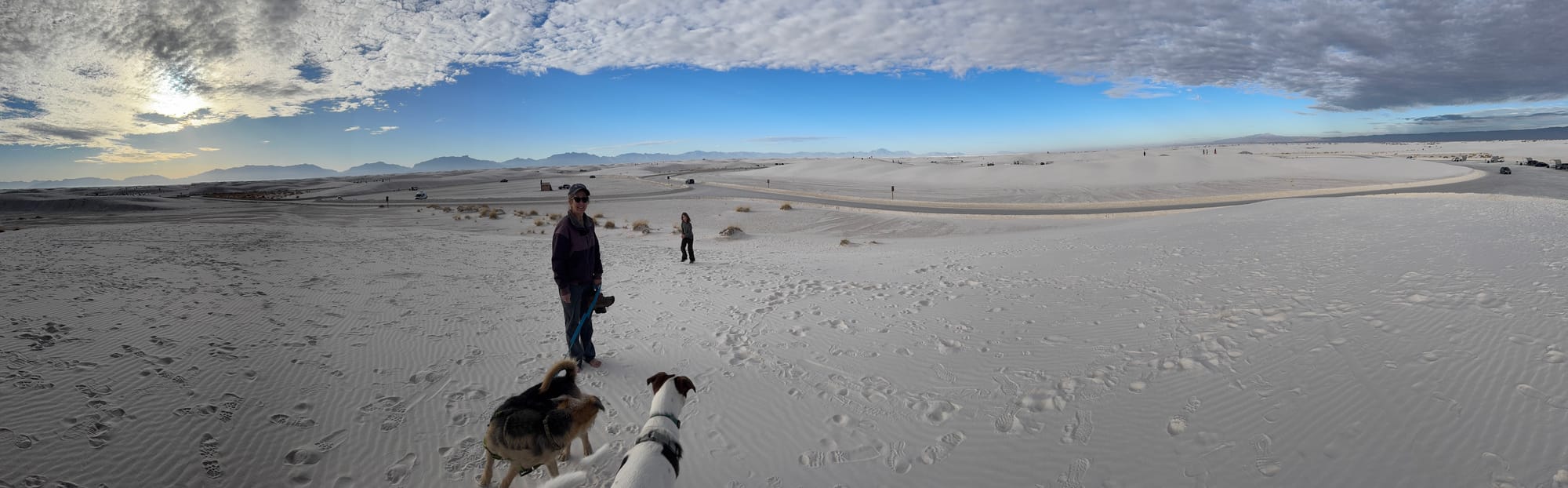 Panorama photo of white sand dunes with a person and two dogs in the foreground, a child further back, and mountains in the distance, under partly cloudy skies.