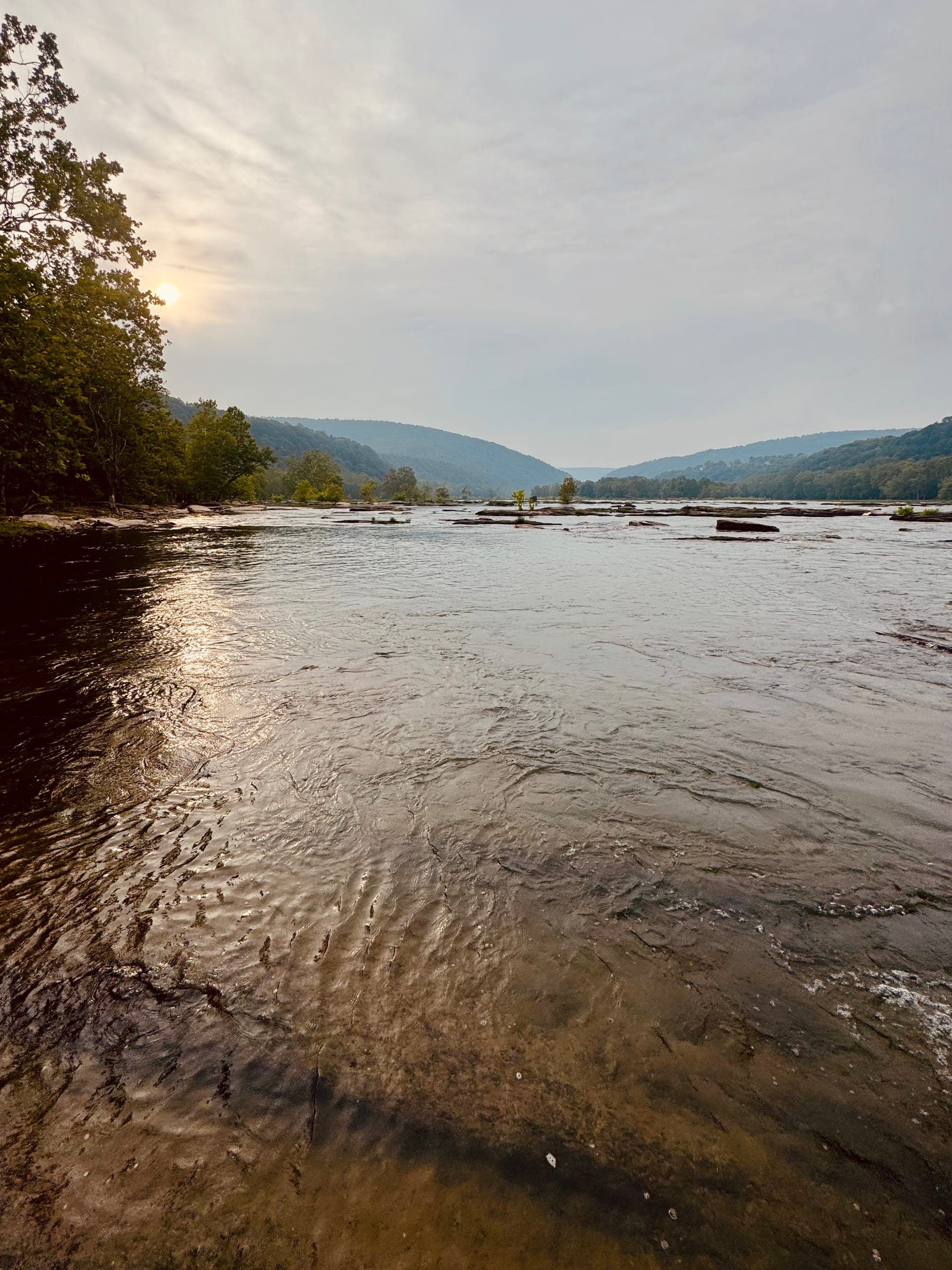 Sun rising through light clouds over mountains in the distance, and with a rocky river in the foreground.