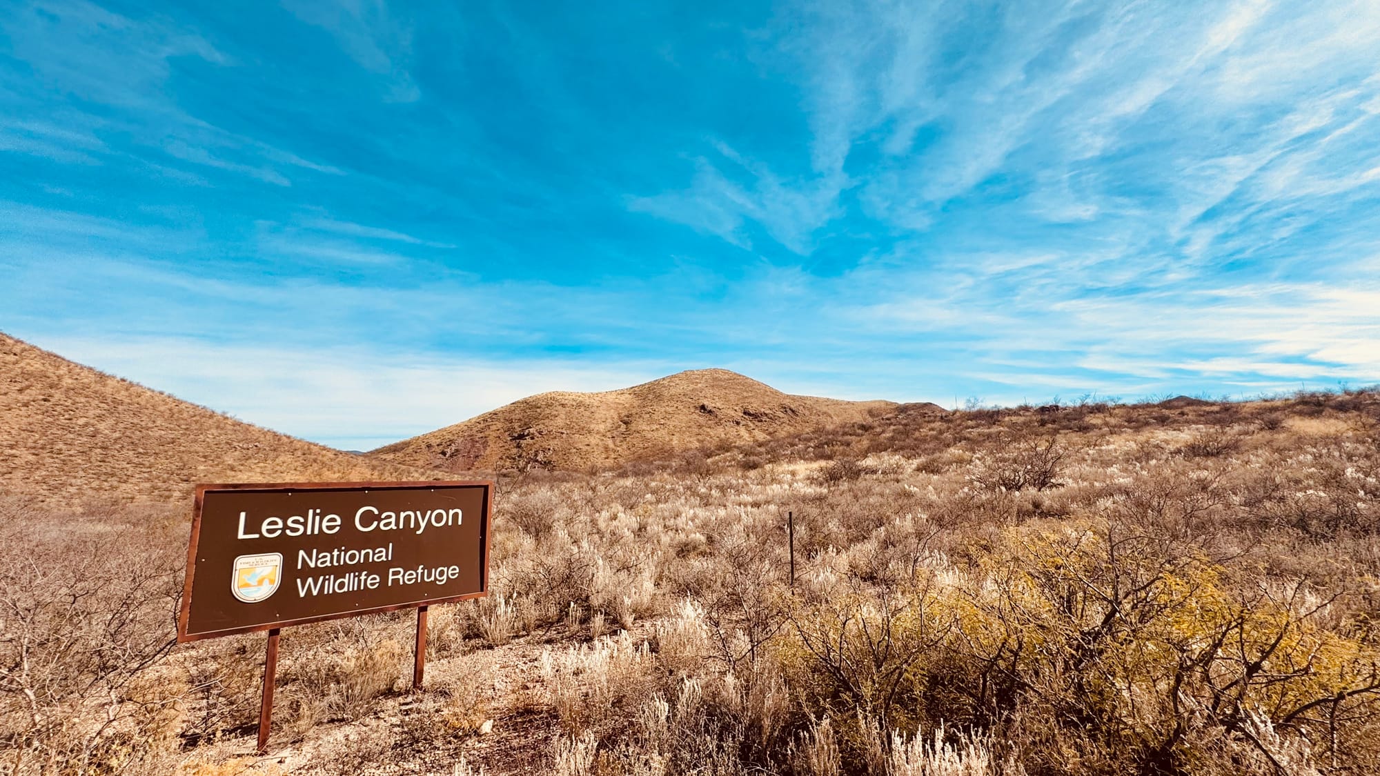 Desert scrub landscape with a sign on the left, and partly cloudy skies overhead.