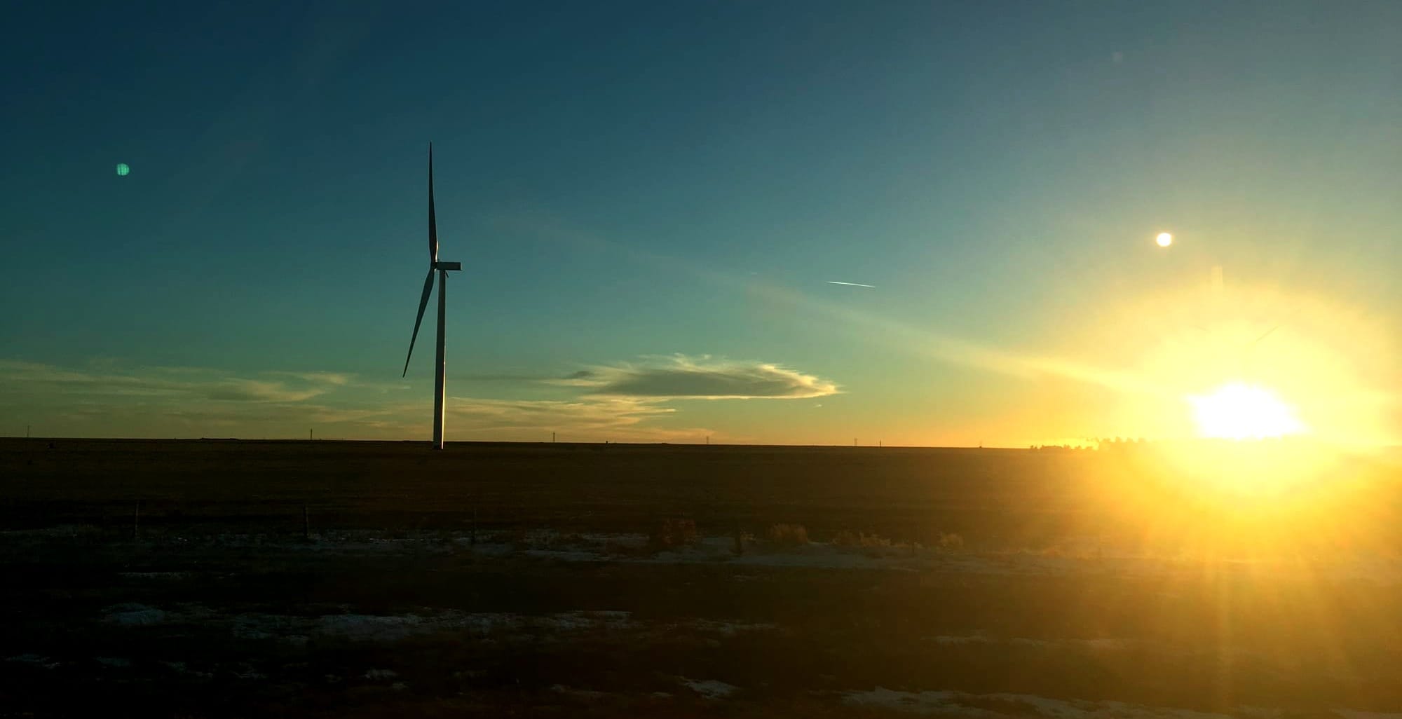 A wind turbine in the foreground with sunset in the background under clear skies.