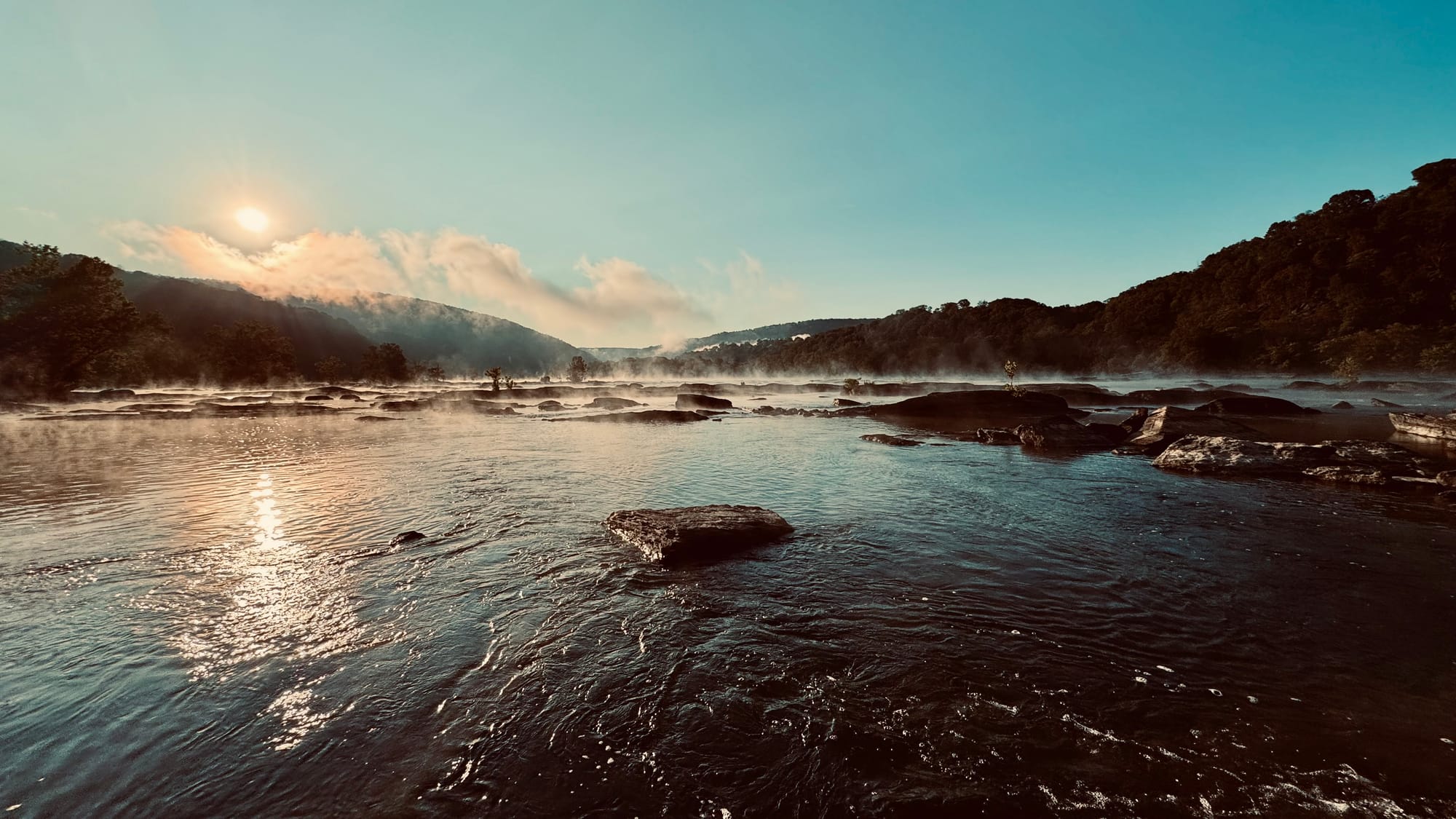 Sun rising in a clear sky with a bit of fog lifting off a river, with hills in the background.