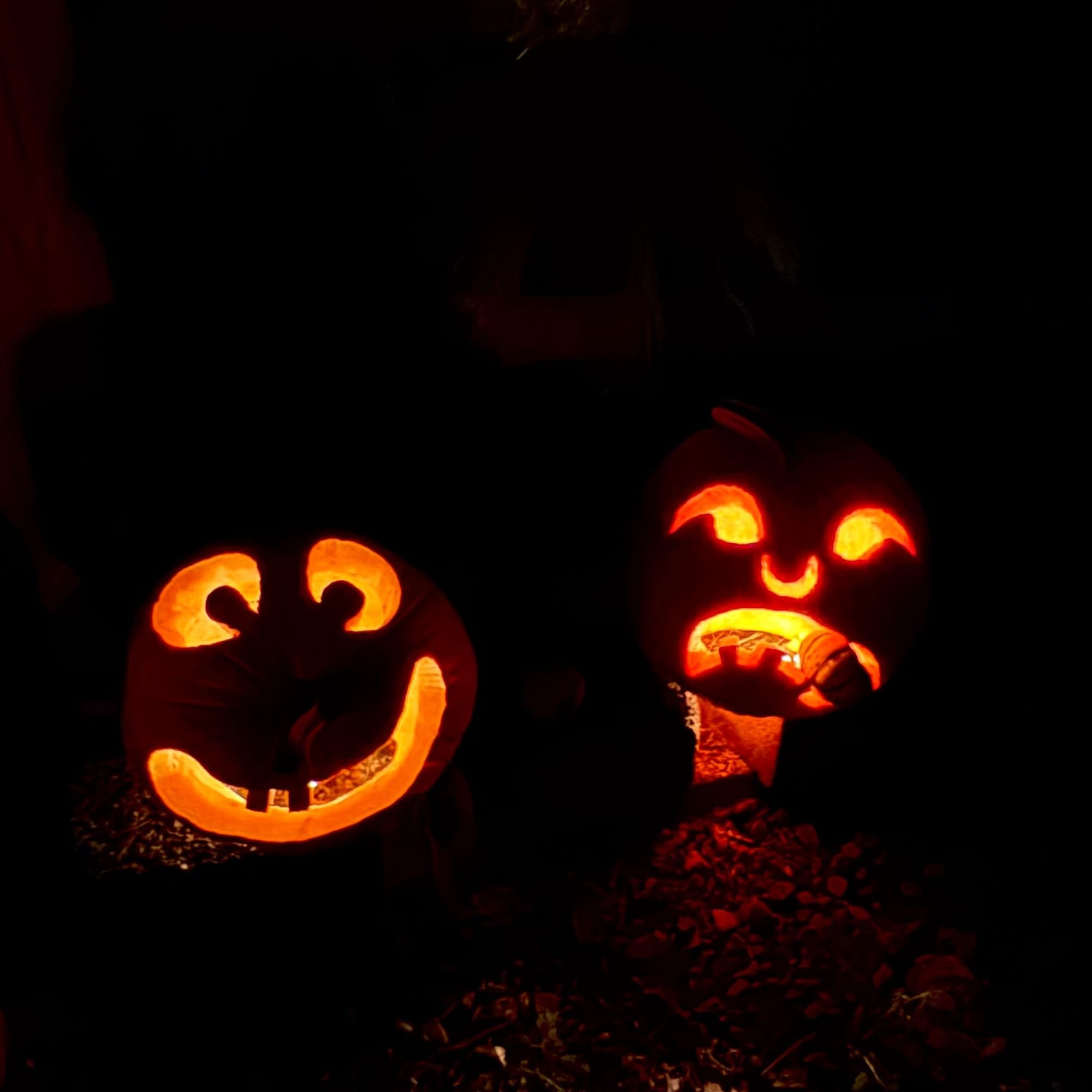 Two jack-o-lanterns, one smiling with buck teeth, one eating a smaller pumpkin, at night and illuminated by candles.