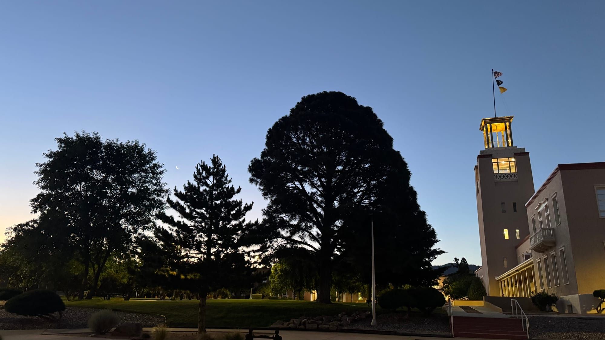 A building with a tower extends into the dark blue, pre-dawn sky, with a few tree silhouettes to the left and a crescent moon above.