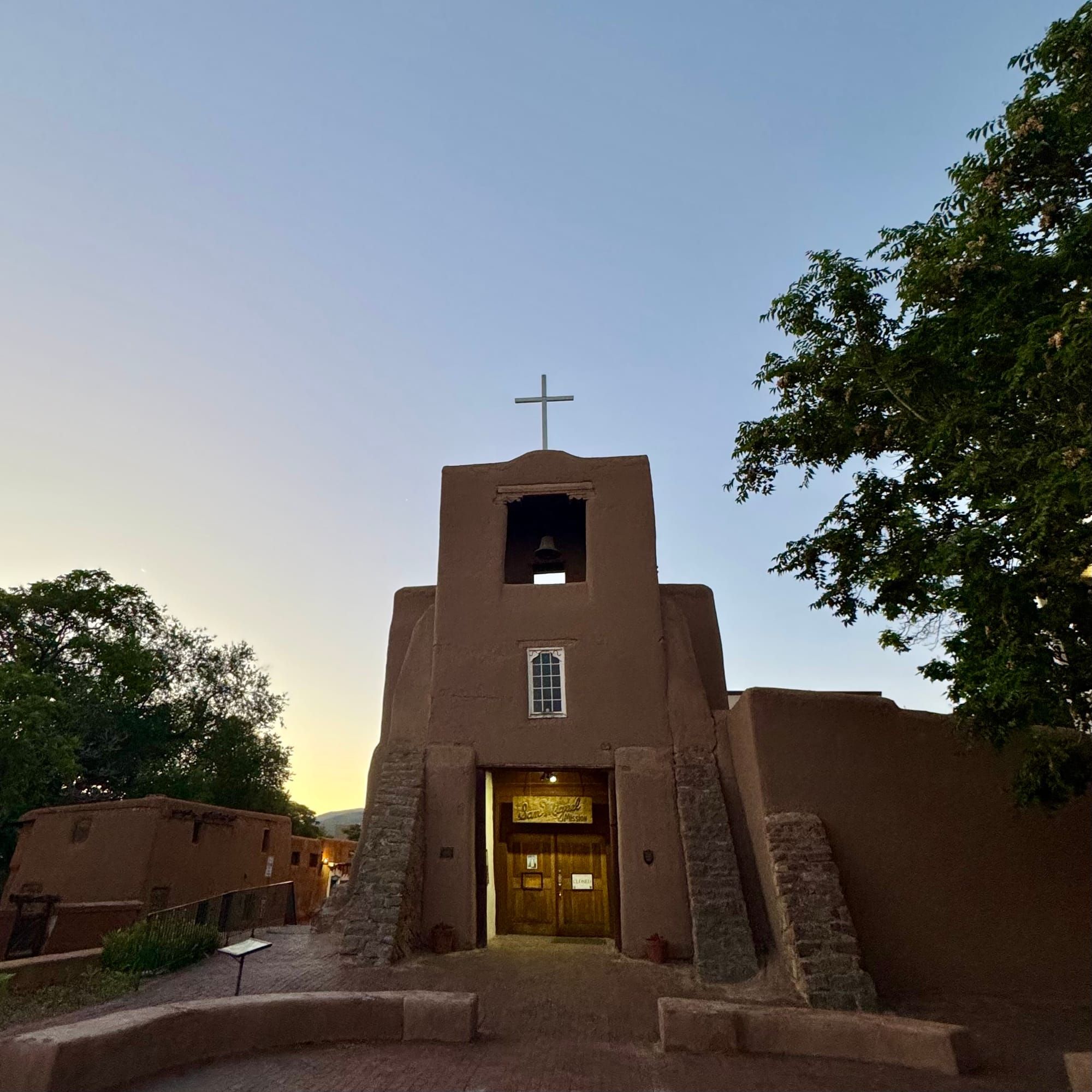 An adobe church tower in the center of the photo with clear skies and in dawn's early light behind, and a few trees surrounding.