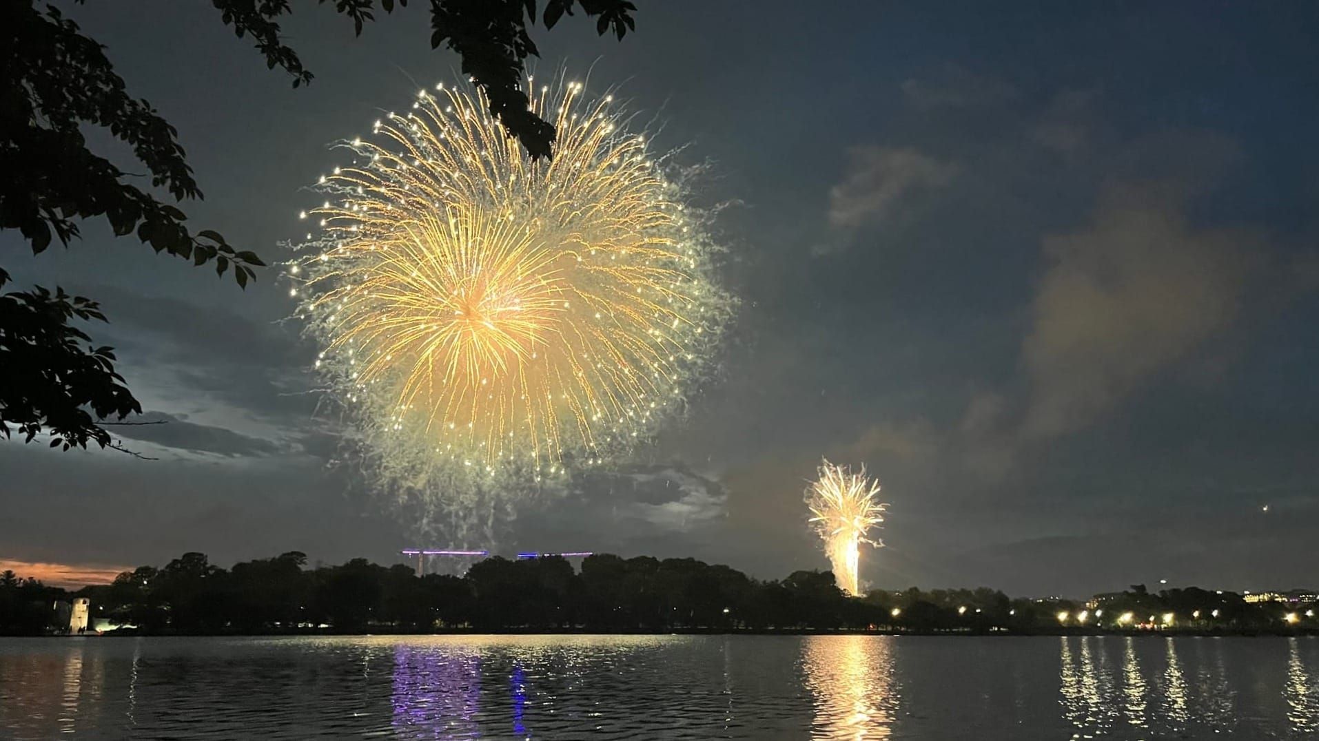 A ball of yellow-orange fireworks explodes against a night sky while a more distain "fountain" of fireworks goes off below. The lights are reflected on water in the foreground.
