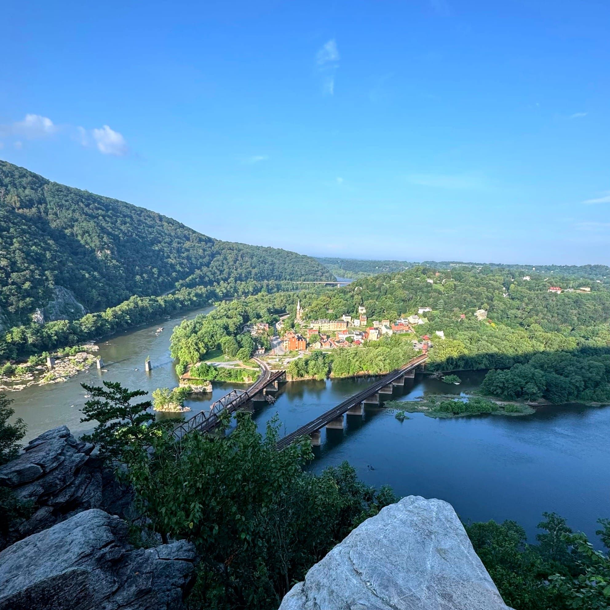 View from a rocky outcrop overlooking to merging rivers, with a small down at the junction, and blues skies with a few clouds overhead.