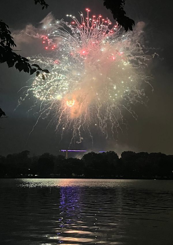 Whitish, red, and green fireworks explode over and reflect in a body of water, with the silhouettes of trees in the distance.