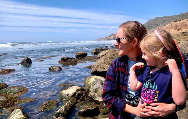 A woman and a girl sit on a rocky shore looking at waves on the ocean, with rocky hills in the background and partly clear skies above.