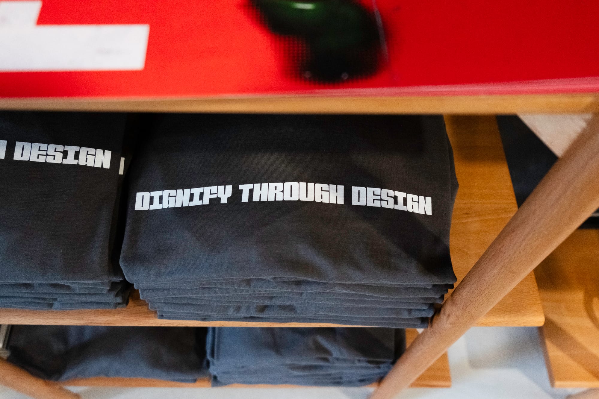 Stacks of t-shirts on a wooden shelf with "DIGNIFY BY DESIGN" on the chest, viewed from above.