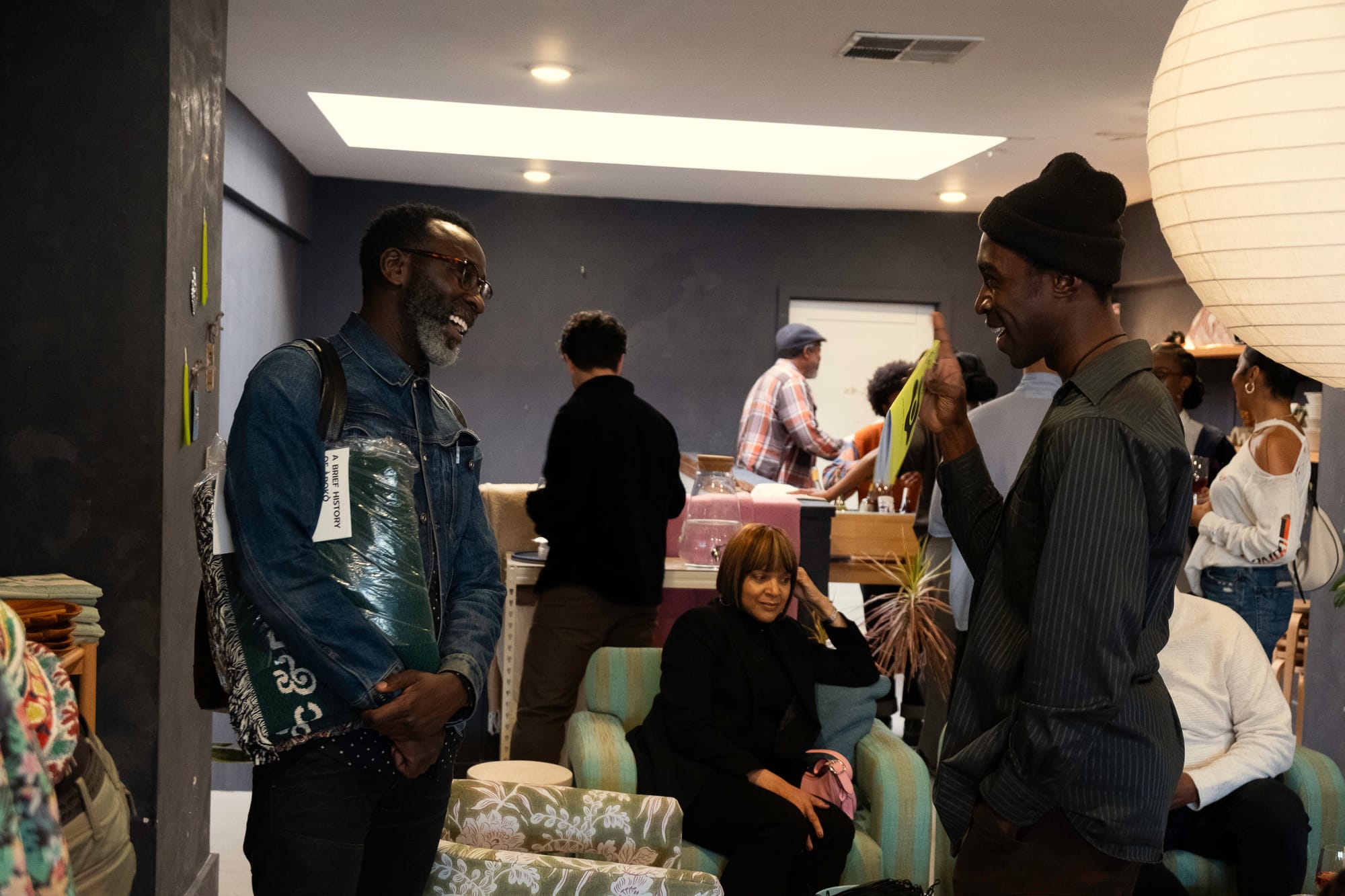 Two Black men face each other in conversation, with other folks milling about in the background.