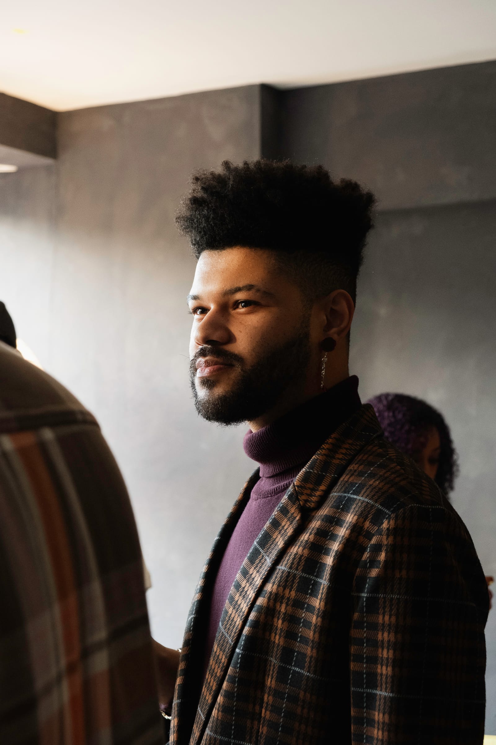 Portrait of a Black person with a high-top textured haircut, beard, and silver hanging earring. They are wearing a maroon turtleneck and brown checkered suit.