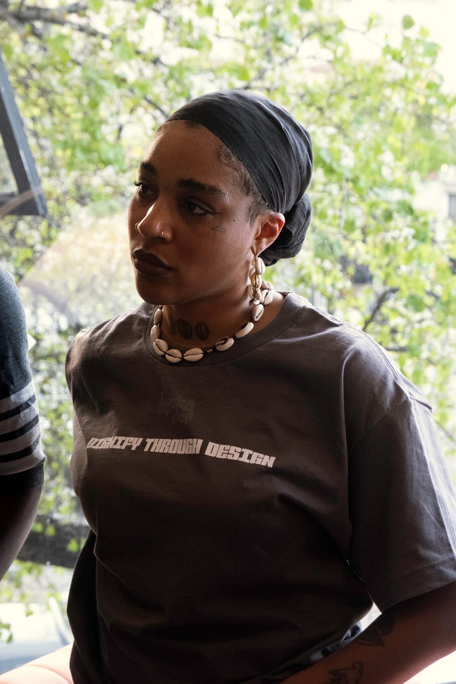 Portrait image of Àrokò member Kristen Stain. They are a Black femme with 2 nose rings, wearing a cowry shell necklace, and a grey t-shirt with the words "DIGNIFY BY DESIGN" on the chest.