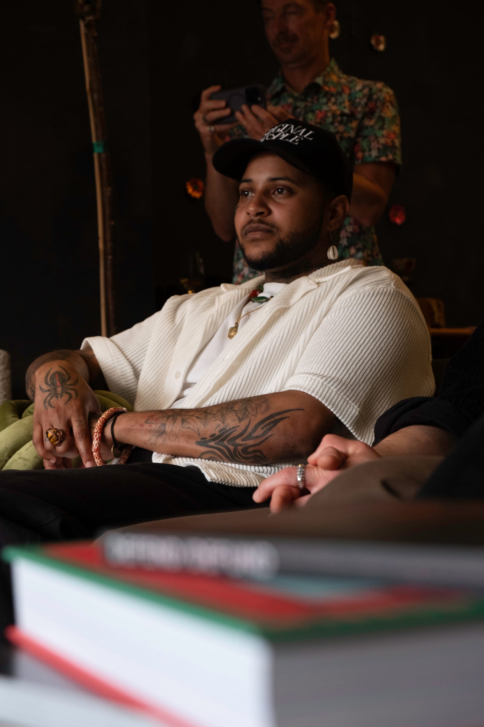 Portrait of a Black person with multiple tattoos, wearing a white, short sleeved collared shirt, and black baseball cap.