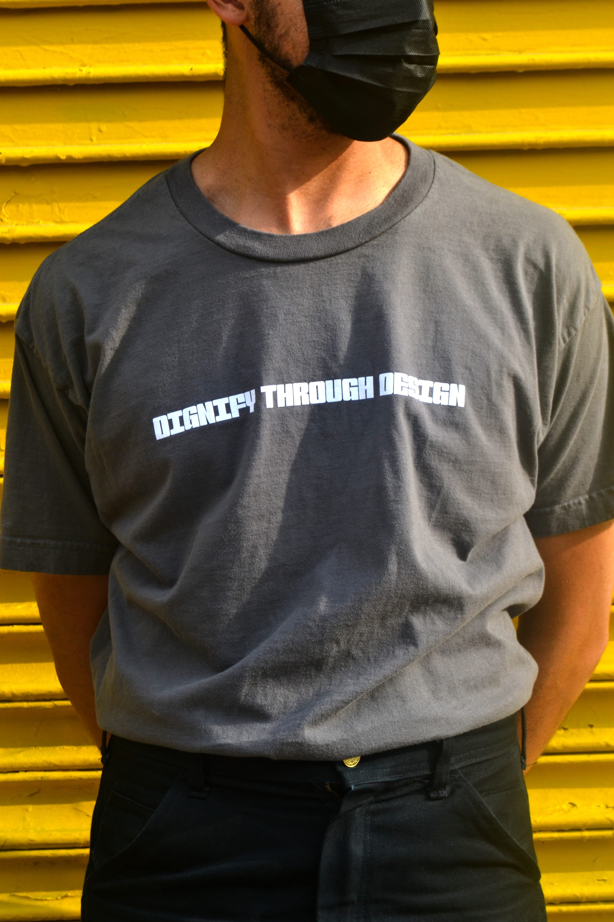 A Black man wearing a mask wears a grey t-shirt with the words 'DIGNIFY THROUGH DESIGN' printed across the chest.