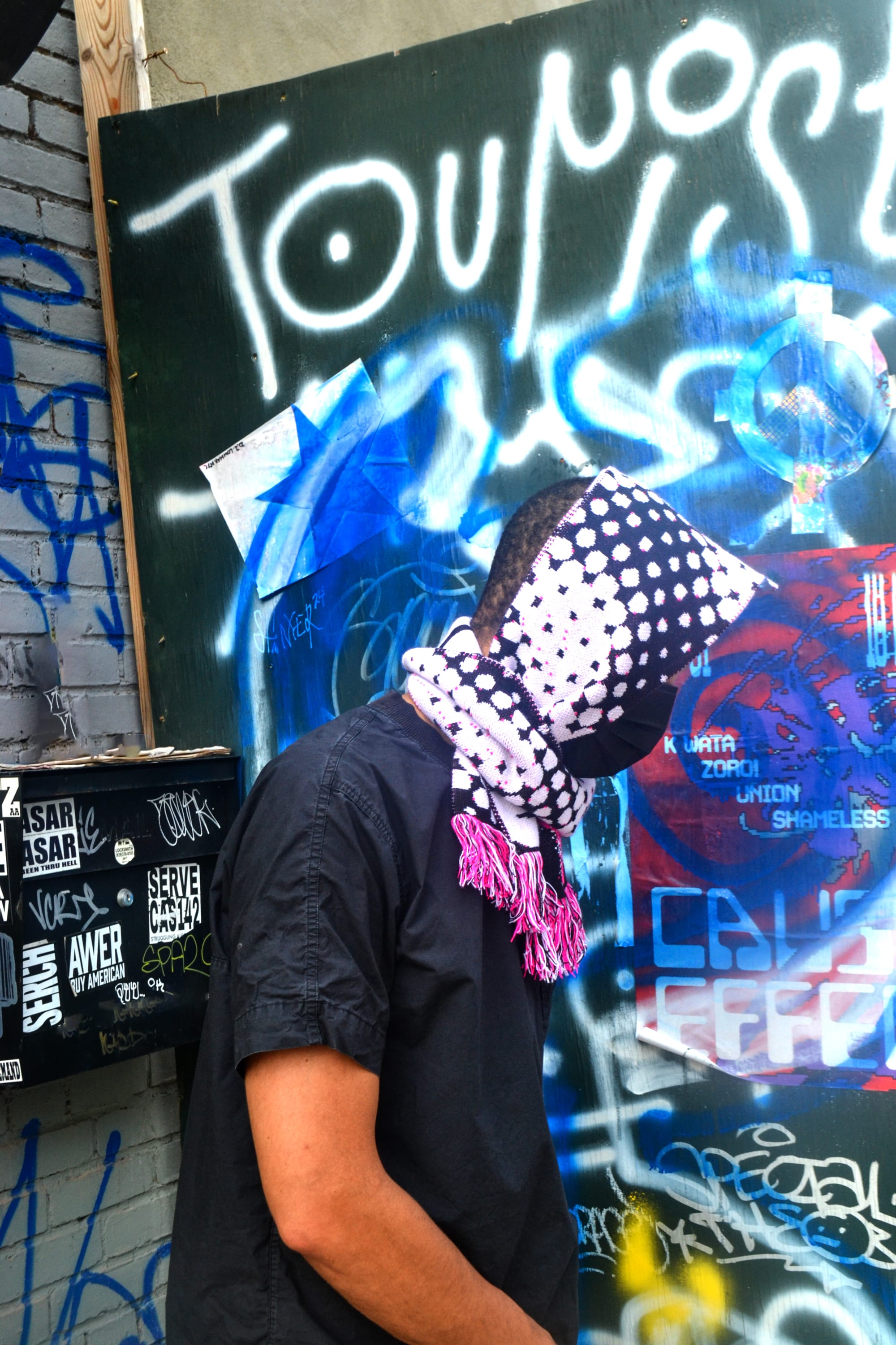 A Black man wears a scarf tied around his head in front of a heavily graffitied construction zone.