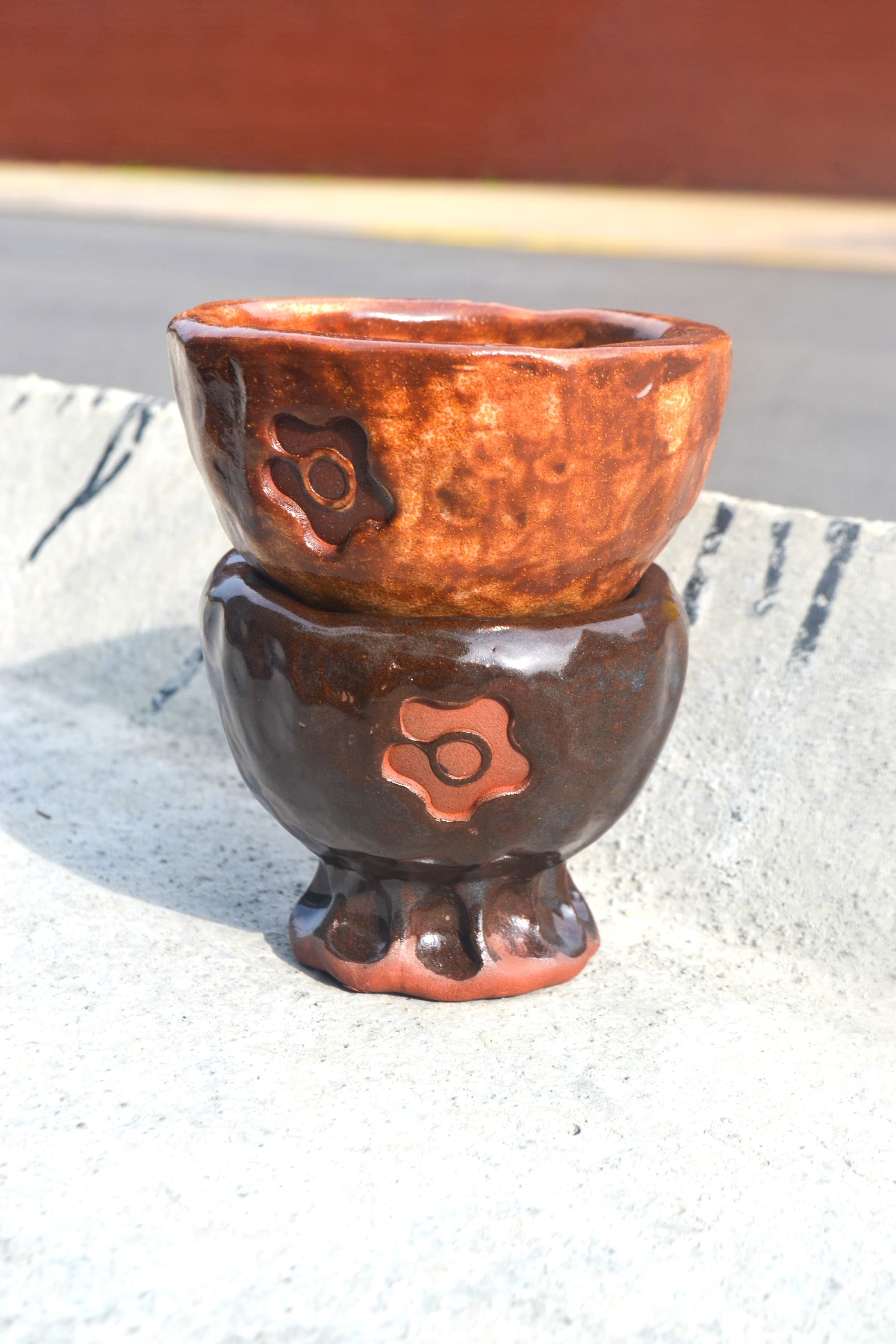 Two brown ceramic cups sit stacked on a concrete road barrier.