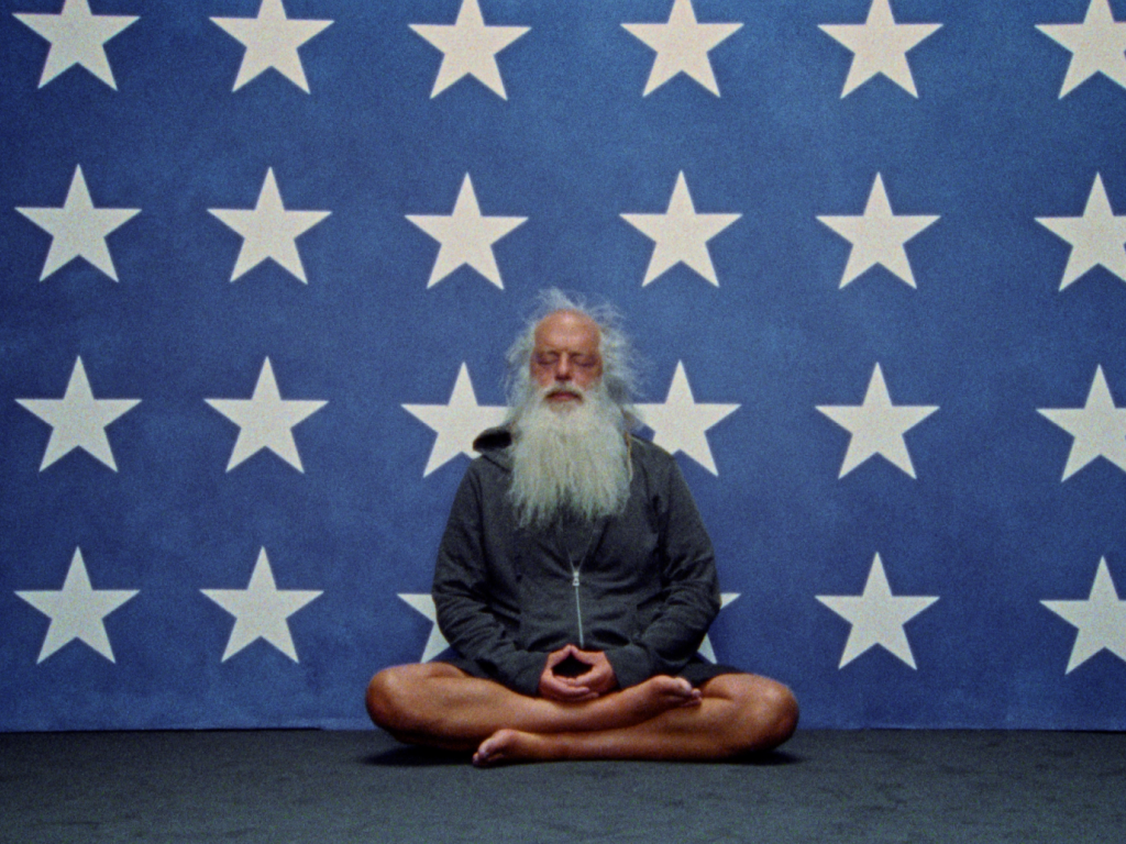 A photo of Rick Rubin fake-meditating in front of the stars portion of an American flag.