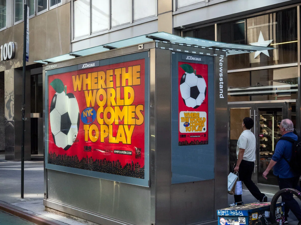 Picture of the 2026 World Cup NYC branding in the wild, with a red poster with a soccer ball / apple that says "WHERE THE WORLD COMES TO PLAY" on a newstand.