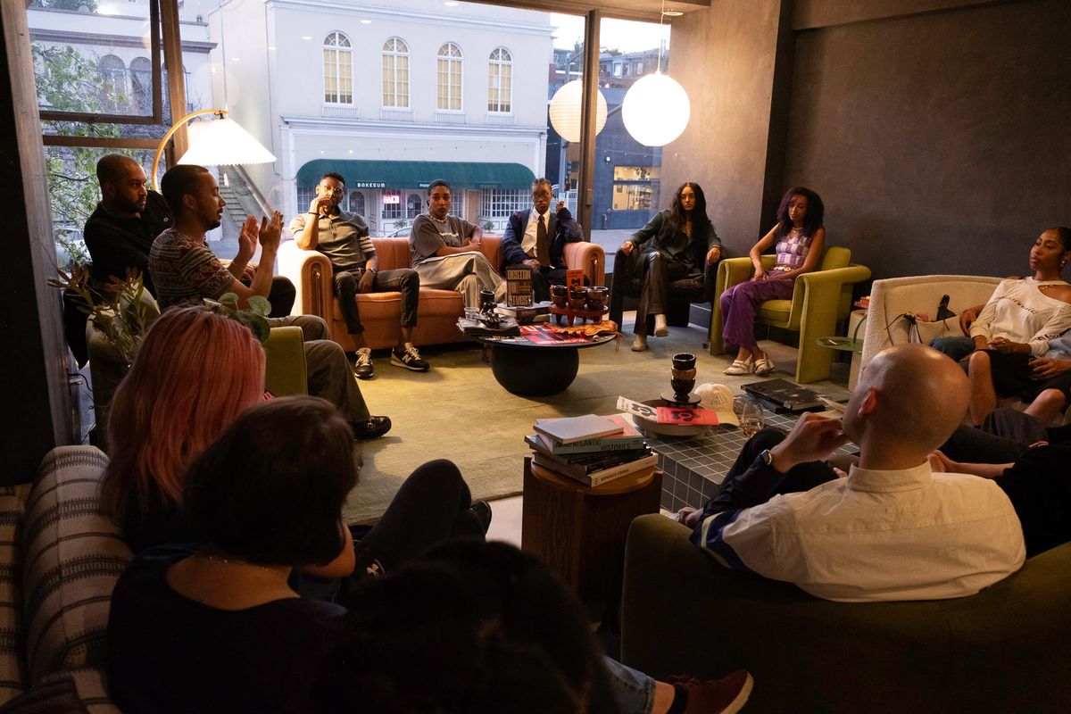 Gathering of seven members of Àròko Cooperative gathered at Alkali Rye in Oakland, CA. They are sitting in a semi-circle with their backs to a full-length window.