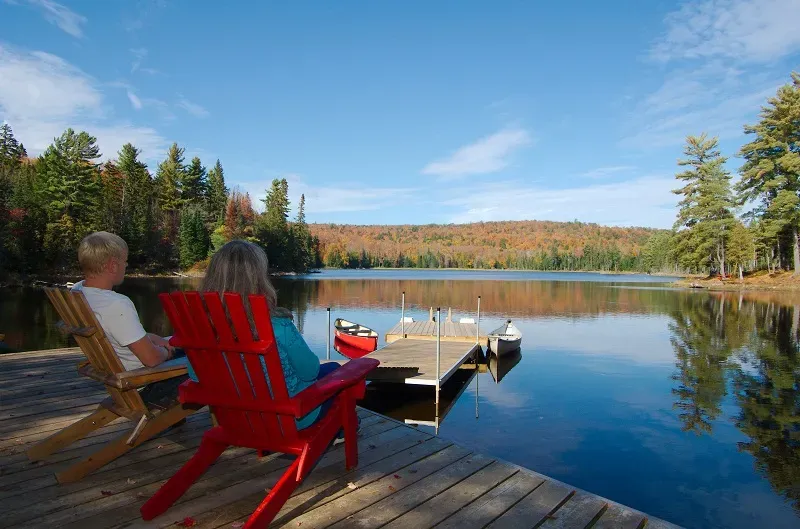 Two guests relax in red Adirondack chairs on a dock overlooking an autumn lake at Algonquin Eco-Lodge, Canada