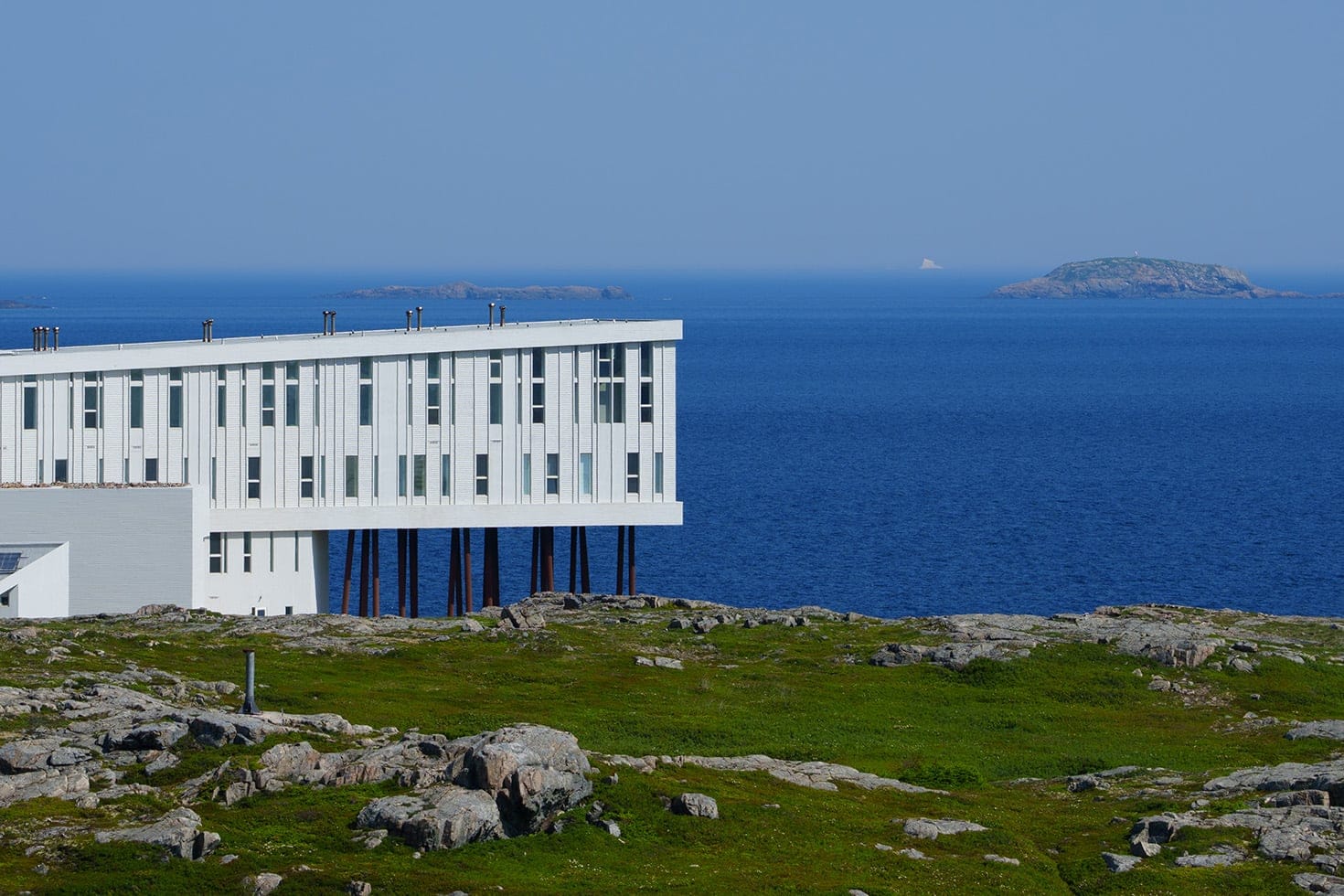 Fogo Island Inn's long white facade raised on rust-red stilts above a rocky, lichen-covered headland, with the deep blue North Atlantic and distant islands beyond, Newfoundland