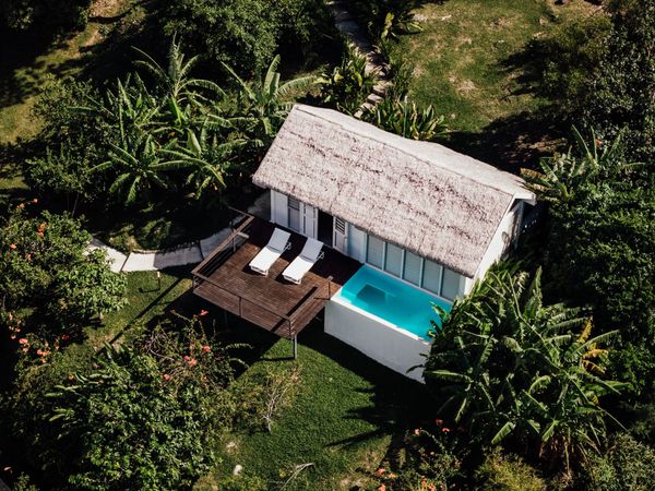 An overhead view of a thatched-roof bungalow with a timber sun deck and small turquoise plunge pool, ringed by banana palms and flowering tropical garden