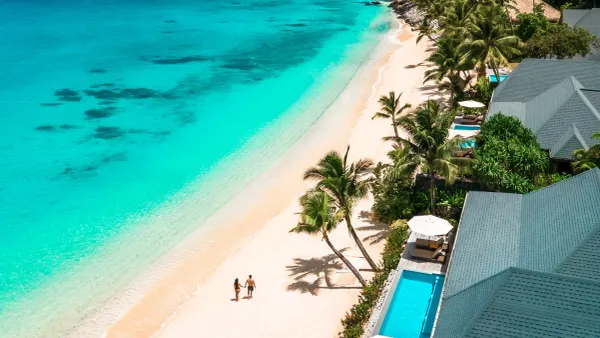 An aerial view of two people walking a white-sand beach beside turquoise shallows, with palm-shaded resort villas and private plunge pools along the shoreline