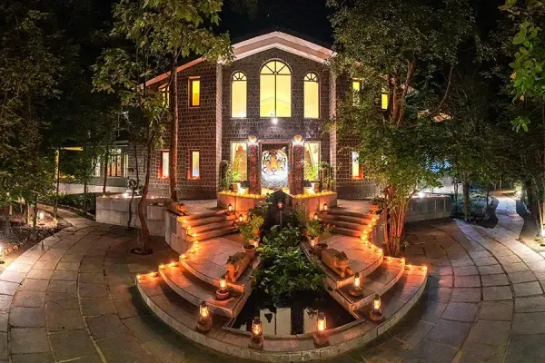 A stone safari lodge lit by lanterns at night, its arched windows glowing warm above curved steps and a tiger mural set into the entrance wall