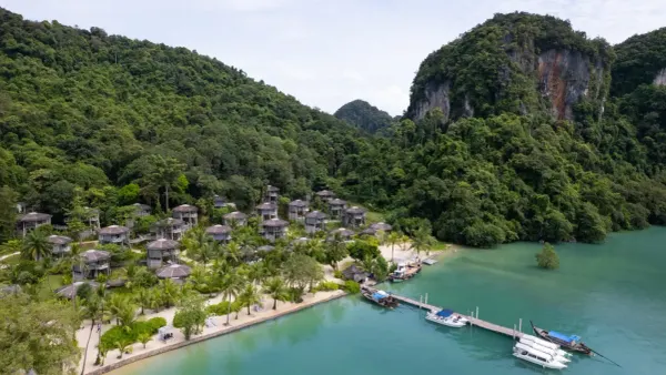 Aerial view of TreeHouse Villas on Koh Yao Noi, with thatched treehouse villas in the jungle above a sand beach, a pier with speedboats and longtail boats, and limestone karsts behind