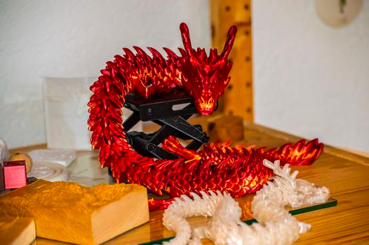 A vibrant red 3D printed articulated dragon posed dramatically on a wooden surface, showcasing its flexible design, with smaller clear dragon models and other objects blurred in the foreground.