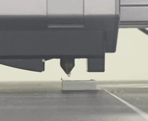 A close-up photo of a 3D printer nozzle positioned above a print bed with a small white object, showing a potential nozzle jam scenario during a printing process.