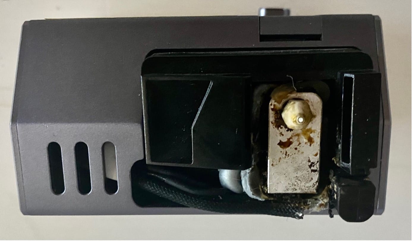 A 3D printer nozzle with visible residue buildup on and around the nozzle tip, showing signs of burnt material and filament leakage. The image emphasizes the importance of regular nozzle cleaning to prevent clogging and heating issues.