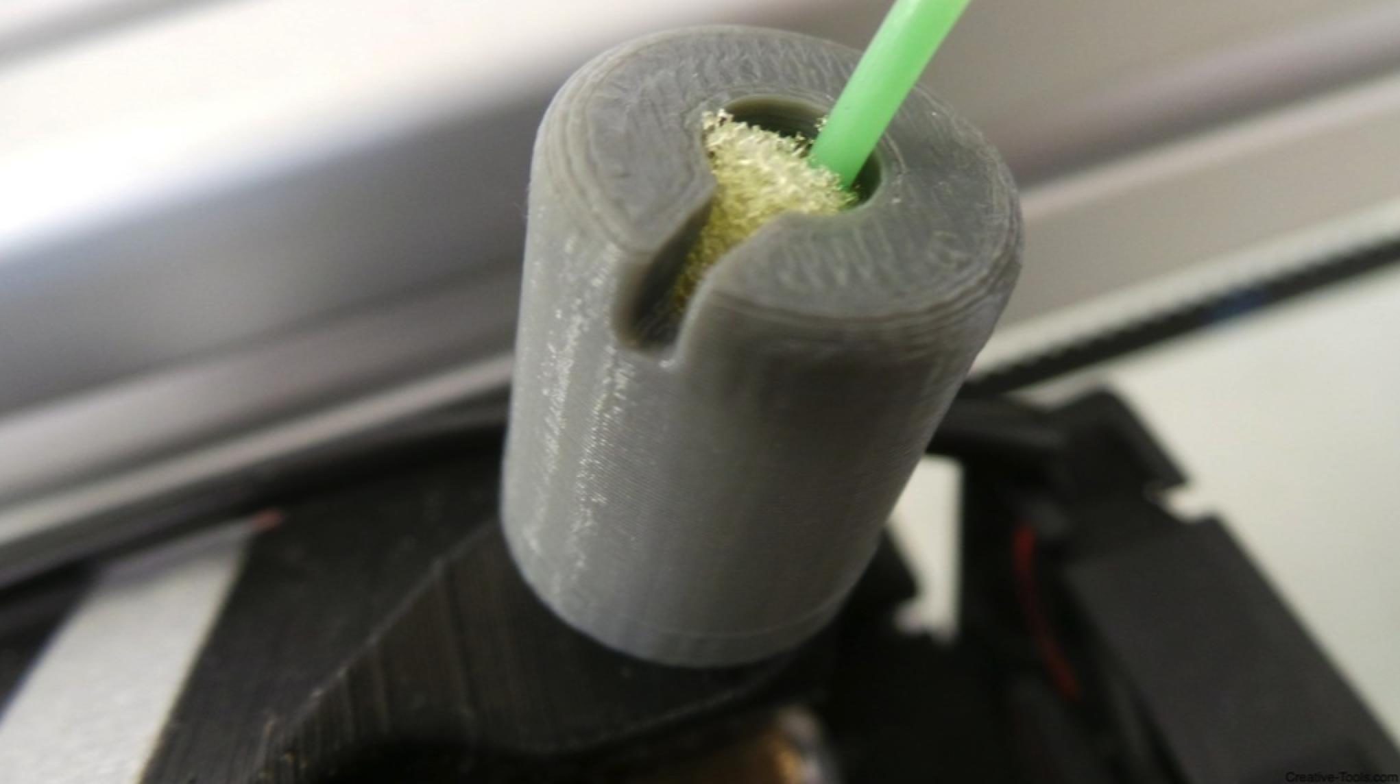 A close-up photo of a 3D-printed gray cylindrical filament guide with a green filament passing through it, containing yellow desiccant inside to absorb moisture, attached to a 3D printer setup.