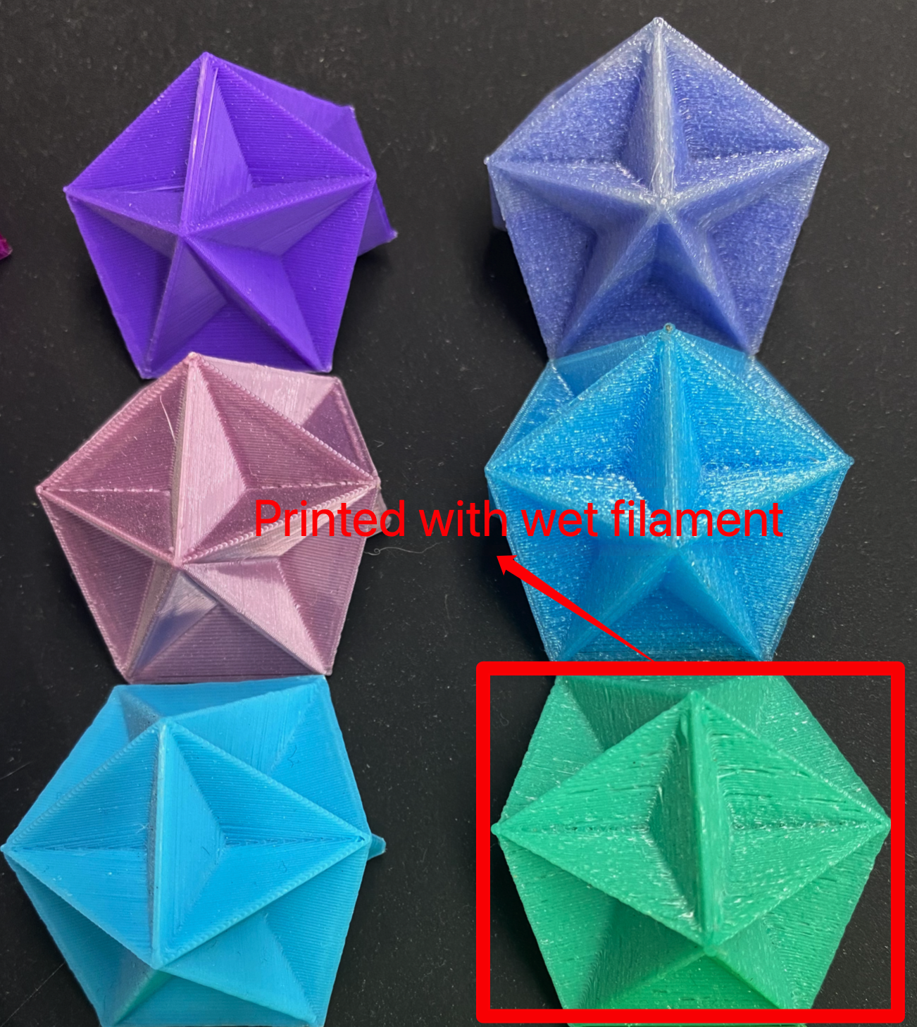 A photo of six 3D-printed polyhedral objects in various colors, with one labeled "Printed with wet filament" in red text, pointing to a blue object with visible stringing and imperfections, while another green object in a red box shows a smoother finish, demonstrating the impact of filament moisture on print quality.