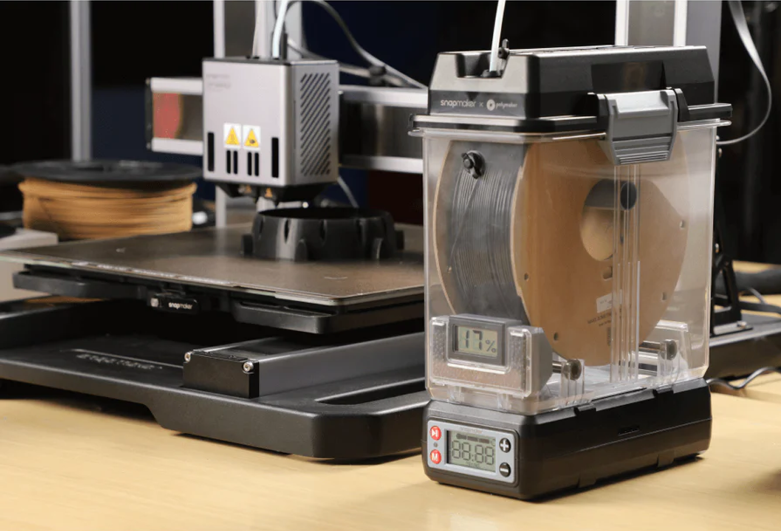Snapmaker filament dryer with a spool of filament inside, positioned beside a Snapmaker 3D printer on a wooden desk. The filament dryer displays a humidity reading of 11.7%.