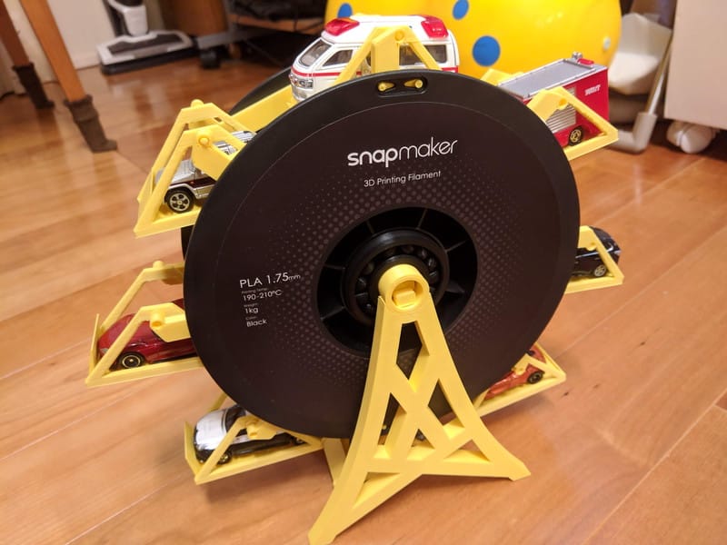A 3D printer filament spool repurposed as a Ferris wheel for toy cars, featuring yellow supports holding various toy vehicles around a Snapmaker PLA spool.