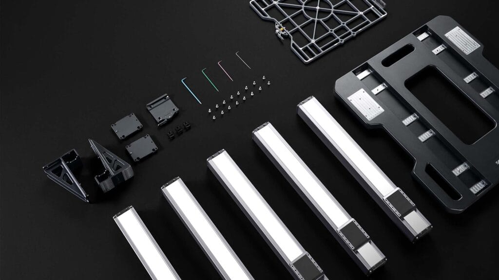 A neatly arranged set of components for assembling a CNC or 3D printer machine, including aluminum extrusions, brackets, screws, hex keys, and structural plates, all laid out on a black background.