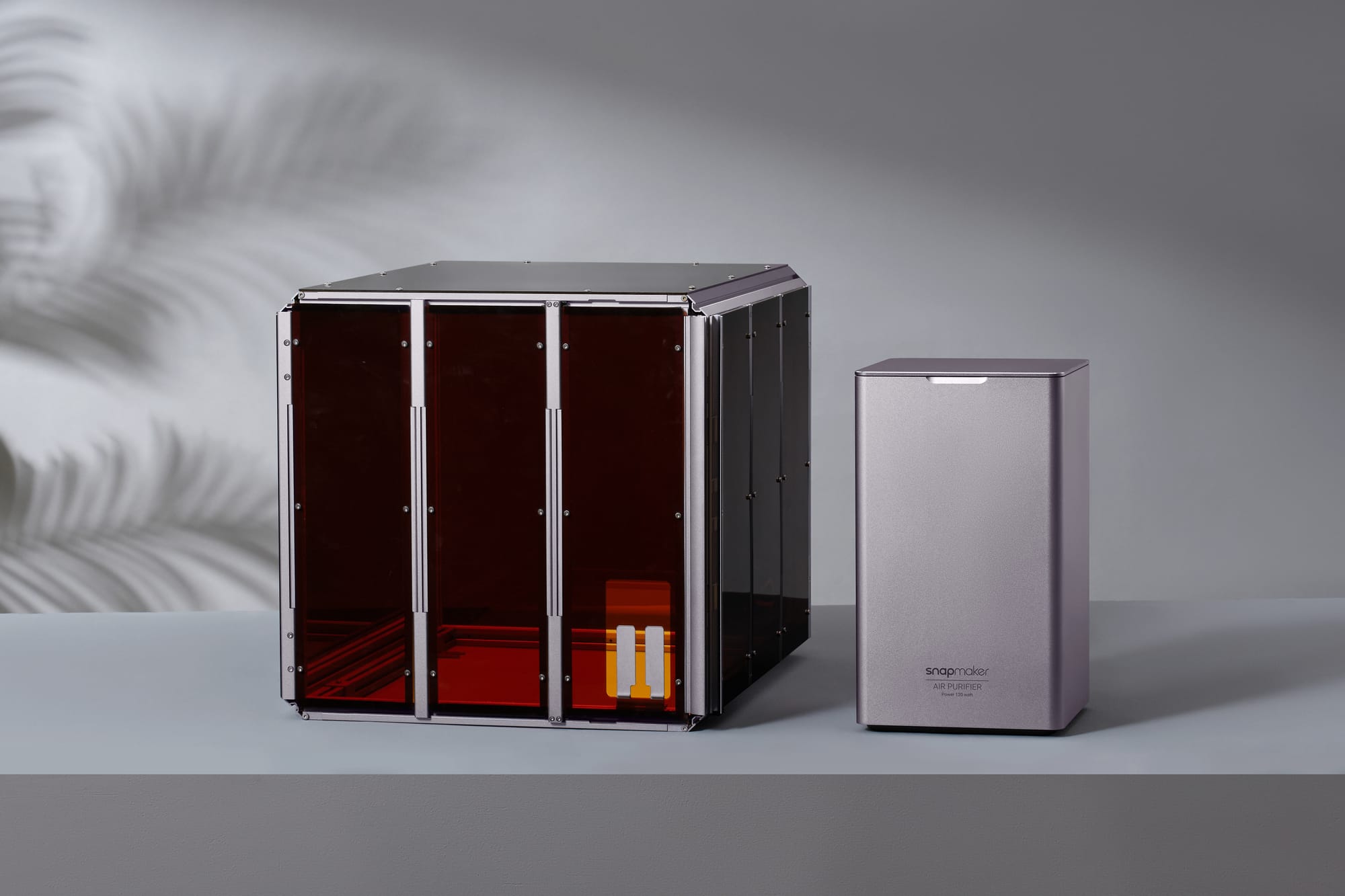 A Snapmaker 3D printer safety enclosure with amber-tinted panels sitting next to a matching standalone air purifier unit on a grey surface.