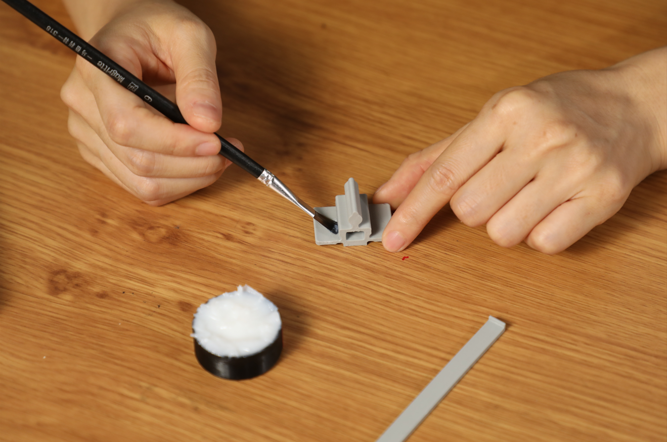 A person uses a small paintbrush to carefully apply adhesive to a small, gray part, demonstrating a technique for precise glue application.