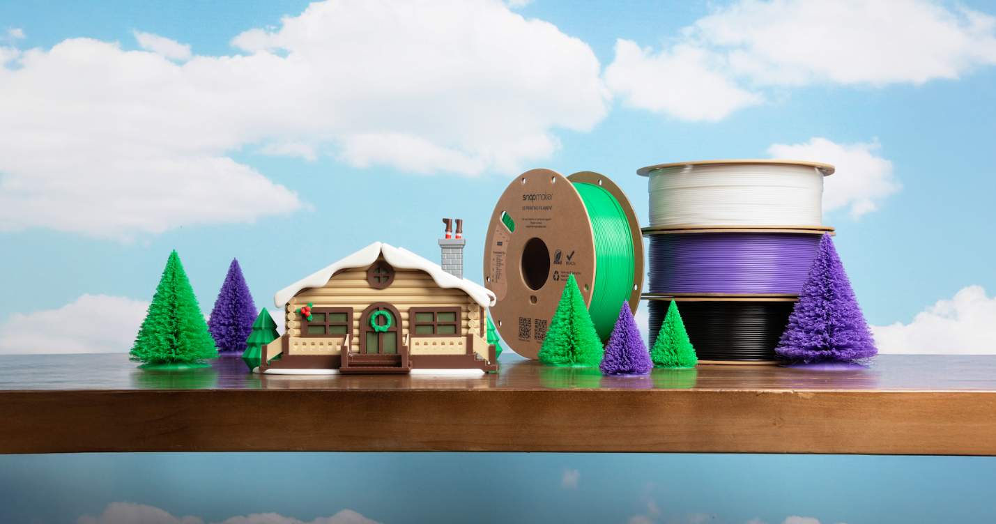 A detailed, 3D-printed winter scene with a log cabin and colorful trees, showcased next to the spools of high-speed PLA filament used to create it.