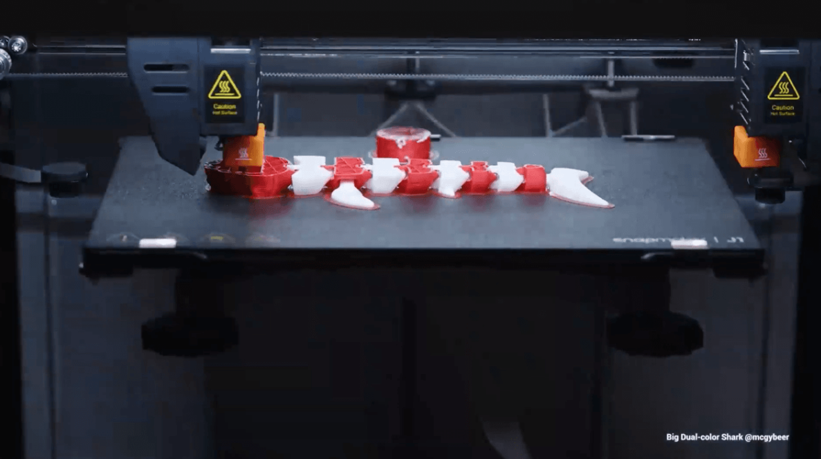 An IDEX 3D printer uses its two independent extruders to print a red-and-white articulated shark, demonstrating the clean color separation of this method.