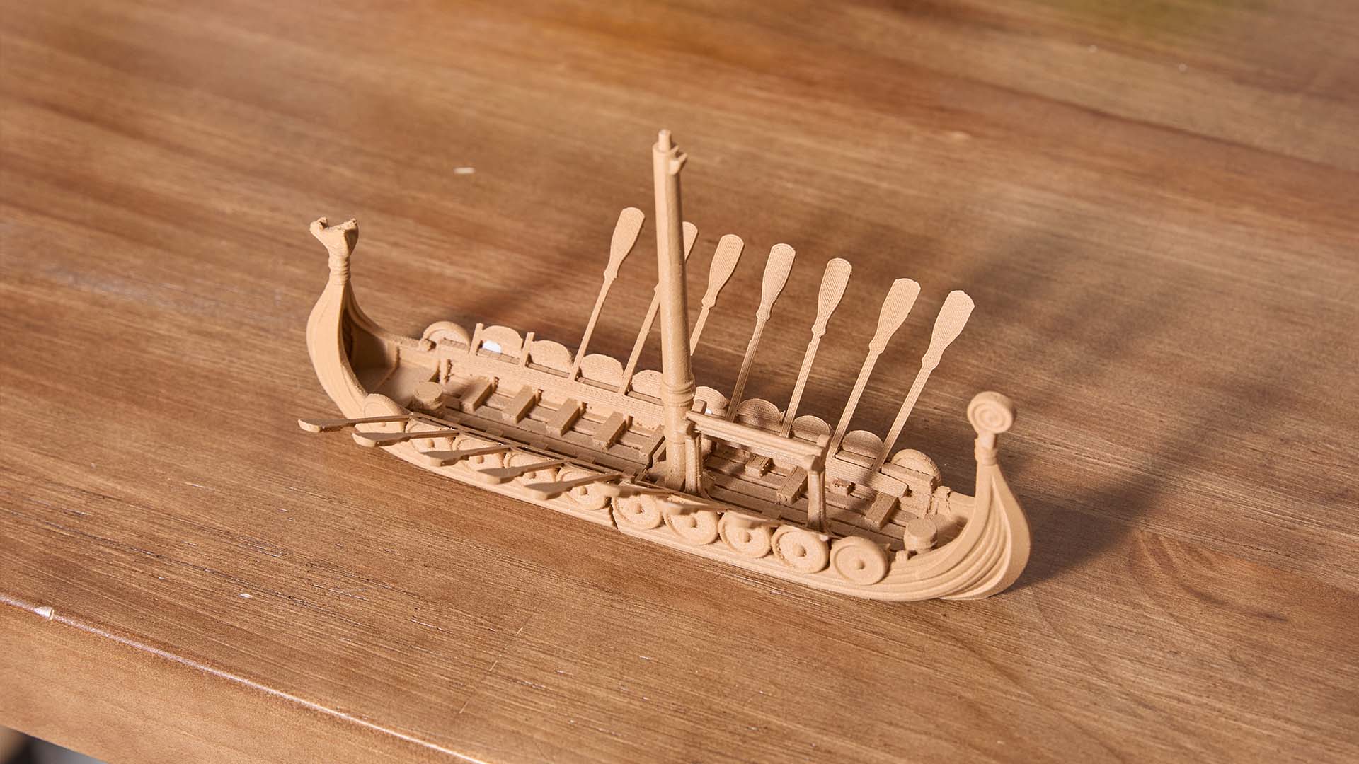  A detailed 3D printed model of a Viking longship made with wood PLA, showing intricate features like oars and shields on a wooden surface.